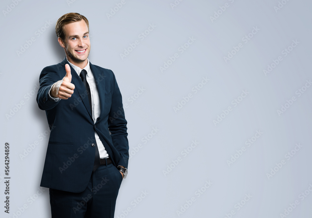 Excited businessman showing thumb up like sign gesture, in black confident style suit, against grey color background. Handsome happy man. Copy space for slogan or text. Excited businessman showing thumb up like sign gesture, in black confident style suit, against grey color background. Handsome happy man. Copy space for slogan or text.