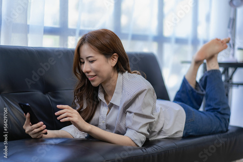 Asian woman holding the phone, looking at the screen, lying on the sofa comfortably At home, happy to relax in the living room, use smartphones, enjoy free time, use social media, surf the Internet. Free Download Asian woman holding the phone, looking at the screen, lying on the sofa comfortably At home, happy to relax in the living room, use smartphones, enjoy free time, use social media, surf the Internet. - GFXLoad