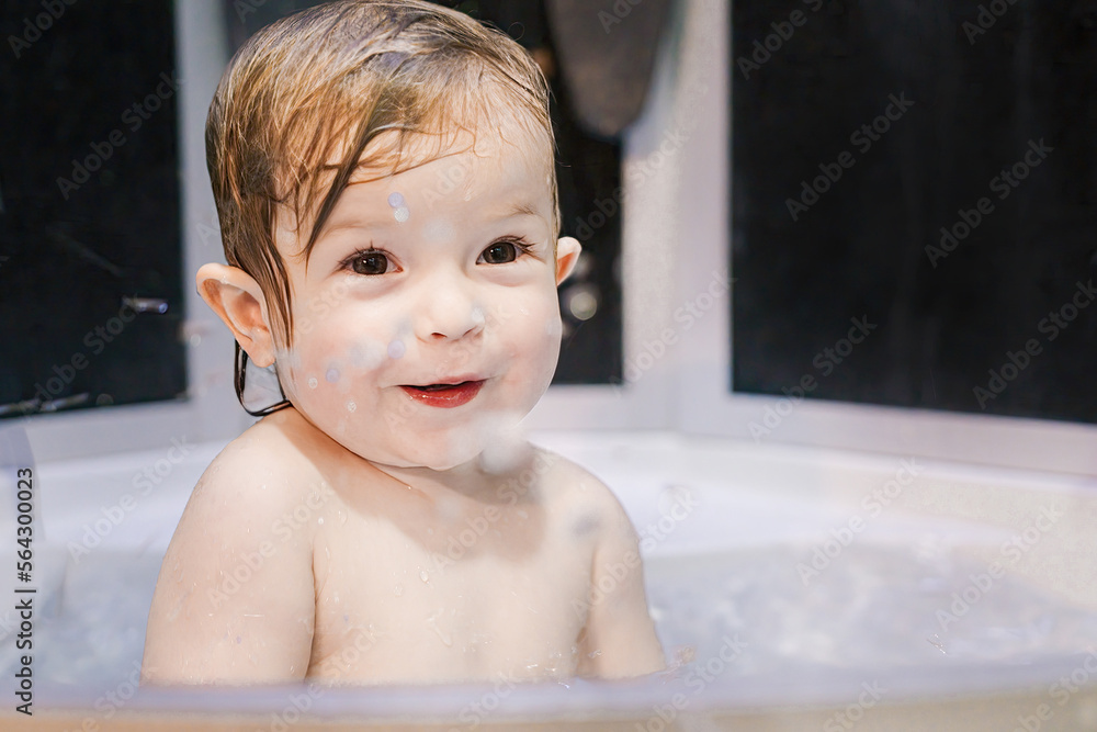 A small child plays in the bathtub and splashes with water. Happy baby with a smiling face and wet hair. Evening hygiene procedures for the baby A small child plays in the bathtub and splashes with water. Happy baby with a smiling face and wet hair. Evening hygiene procedures for the baby