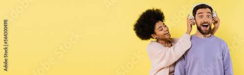 curly african american woman putting wireless headphones on amazed man isolated on yellow, banner.
