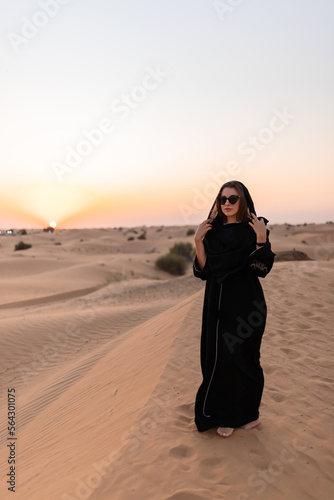 Beautiful mysterious woman in traditional arabic black long dress stands in the desert on sunset