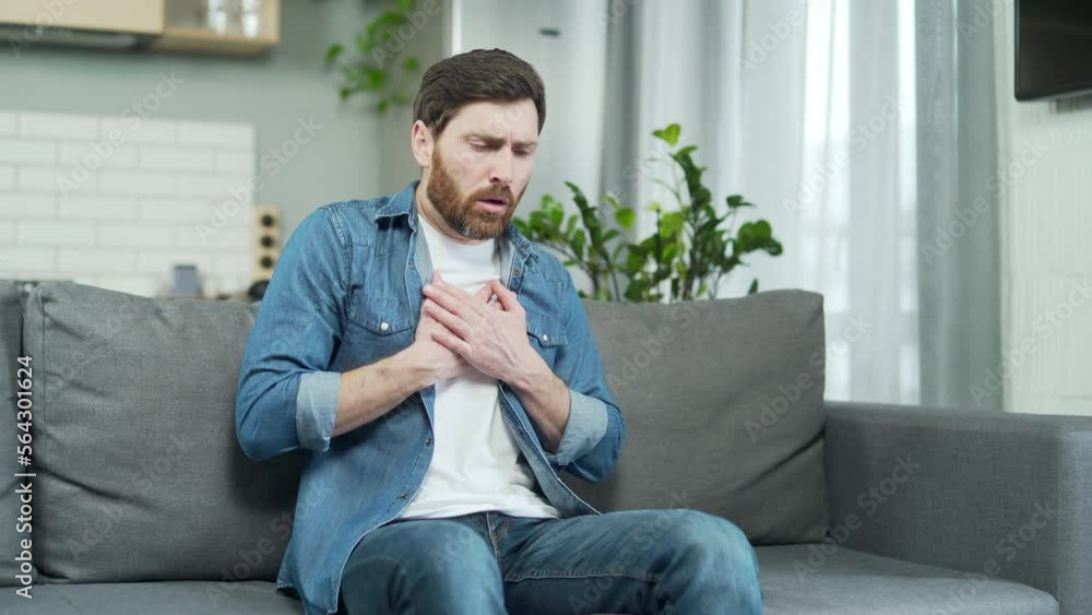 Man having heart attack sitting alone at home on the sofa. Mature bearded Caucasian male holding chest and breathing heavily in room. Indoor Suffering from chest pain on sofa Man having heart attack sitting alone at home on the sofa. Mature bearded Caucasian male holding chest and breathing heavily in room. Indoor Suffering from chest pain on sofa