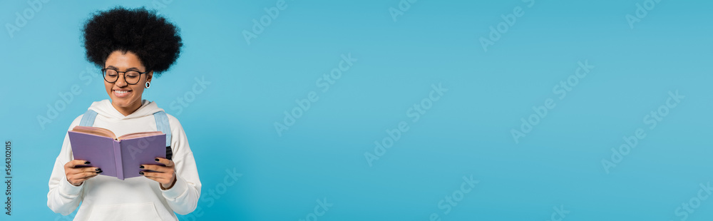 happy african american student in trendy hoodie and eyeglasses reading book isolated on blue, banner. happy african american student in trendy hoodie and eyeglasses reading book isolated on blue, banner.