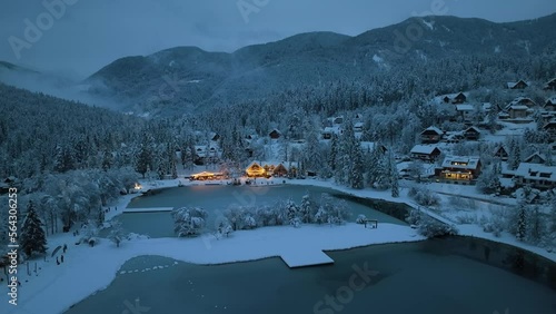 Aerial view of fairy forest in snow, frozen lake and house with lights at night in winter. Top view from drone of mountain village, pond, illumination, snowy pine trees, low clouds at dusk in Slovenia