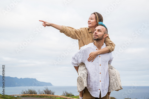 A young couple walks on a road surrounded by nature, with the man carrying the woman on his back