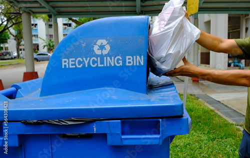 Photography Hands holding white trash bag of recyclable items materials; putting it into a blue recycling bin