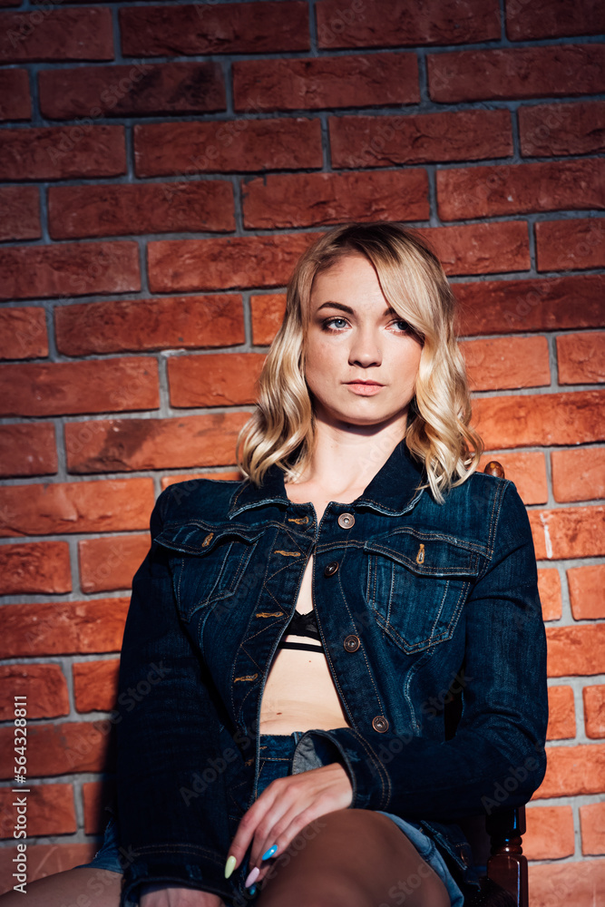 a blonde woman in dark clothes sits on a chair against a brick wall