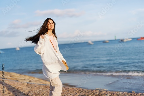 A woman happy in the sun walking on the beach with her arms spread out against the ocean with a toothy smile, flying hair, tanned skin, relaxation, a trip to the ocean and freedom, sunset light.