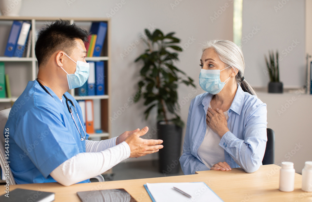Obraz premium Serious mature korean doctor in protective mask consults old european female patient in clinic