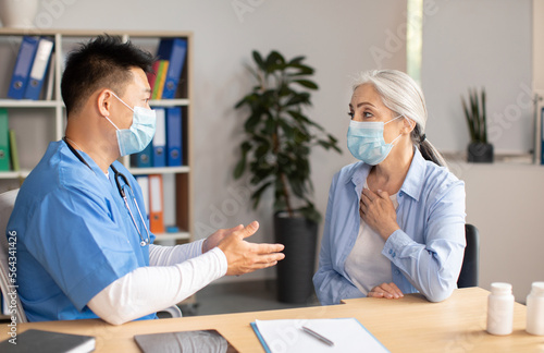Serious mature korean doctor in protective mask consults old european female patient in clinic