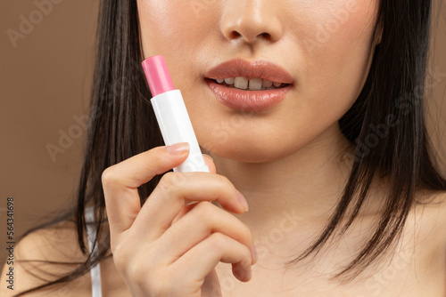 Cropped image of young asian lady holding pink lipstick in hand near face  brown background  studio shot  closeup