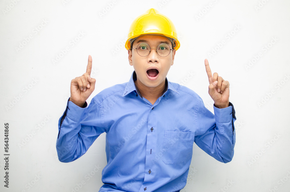 A happy, shocked asian consrtuction worker in blue shirt and yellow safety helmet pointing finger and presenting something ads. 