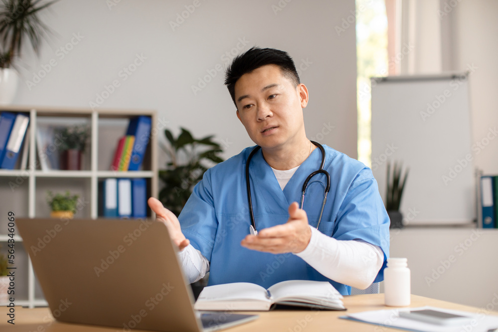 Serious mature chinese therapist in uniform look at computer, talking, gesturing at table in clinic Serious mature chinese therapist in uniform look at computer, talking, gesturing at table in clinic