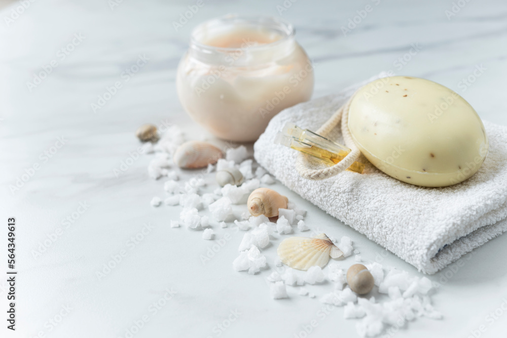 Soap and vials of oil on the white towel, skin cream and scattered sea salt and shells on the light marble background. Copy space