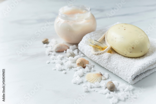Soap and vials of oil on the white towel, skin cream and scattered sea salt and shells on the light marble background. Copy space