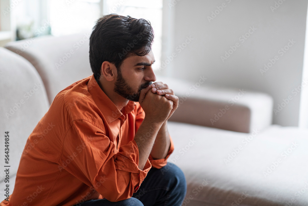 Frustrated arab guy sitting on sofa at home, side view