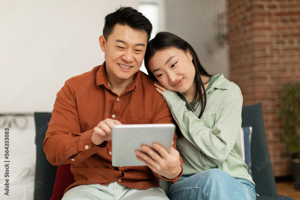 Happy asian spouses using digital tablet, watching videos or surfing internet, relaxing on couch in living room Happy asian spouses using digital tablet, watching videos or surfing internet, relaxing on couch in living room