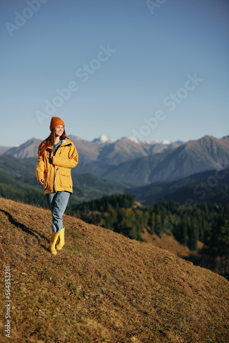Woman walking full-length on a hill in the autumn and looking out at the mountains in a yellow raincoat and jeans happy camping trip, freedom lifestyle 