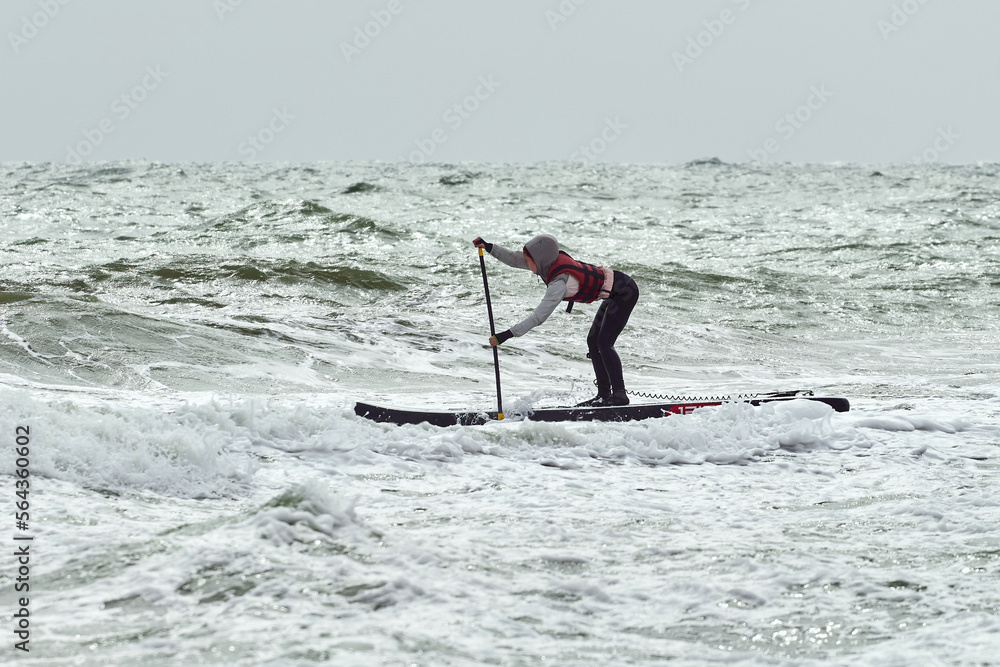 Naklejka premium Young courageous athlete woman surfing by wave on stand up paddle board and catching waves at winter period in sea. Splashes of water. Sea waves, landscape, water sports