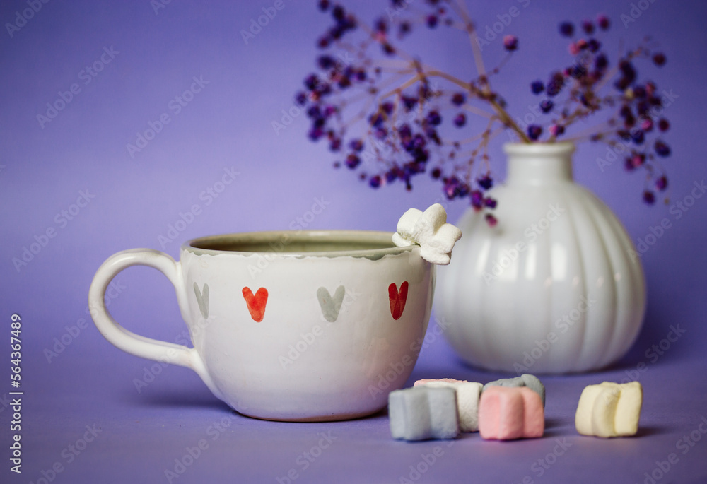Still life white ceramic coffee cocoa tea cup on table on cozy violet background, bouquet of flowers in vase. Symbols of love - hearts on mug. Sweets and treats. Marshmallow assortment Valentines day