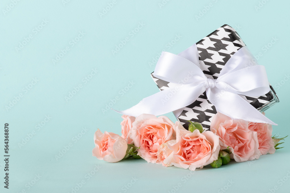 Fototapeta premium Gift box in black and white with a white bow and a bouquet of roses on a blue background.