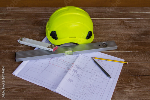 Image of a work table where plans for a building site are placed, a work helmet, a level, tape measure and pencils. Construction engineer job

