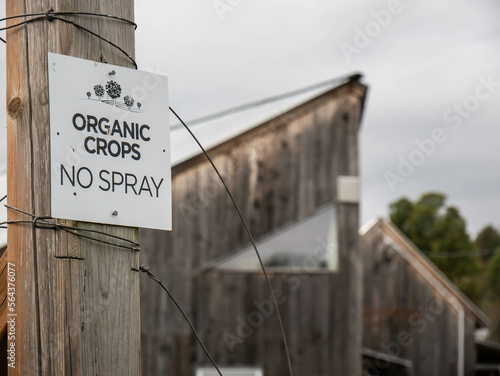 Sign on a wooden fence displaying Organic Crops No Spray with a wooden barn in the background