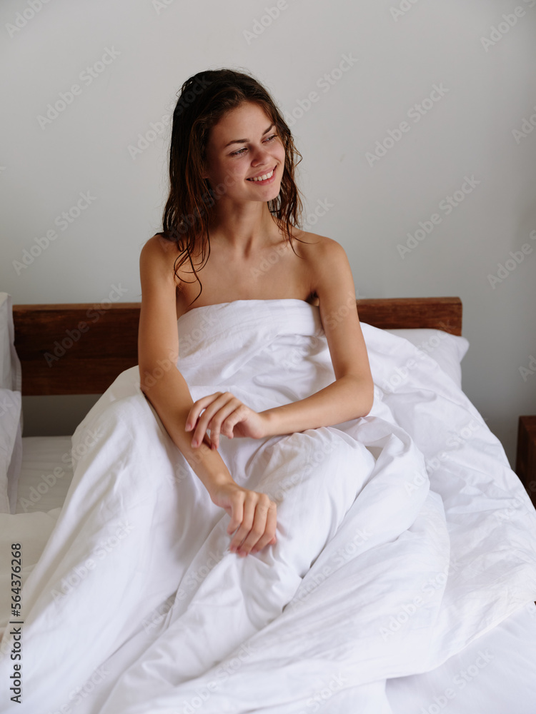 A woman sits on a bed in bedroom and covers her body with a blanket, early awakening, sleepy look, spread her arms and stretches from sleep, happy look, healthy sleep on an orthopedic mattress, smile A woman sits on a bed in bedroom and covers her body with a blanket, early awakening, sleepy look, spread her arms and stretches from sleep, happy look, healthy sleep on an orthopedic mattress, smile