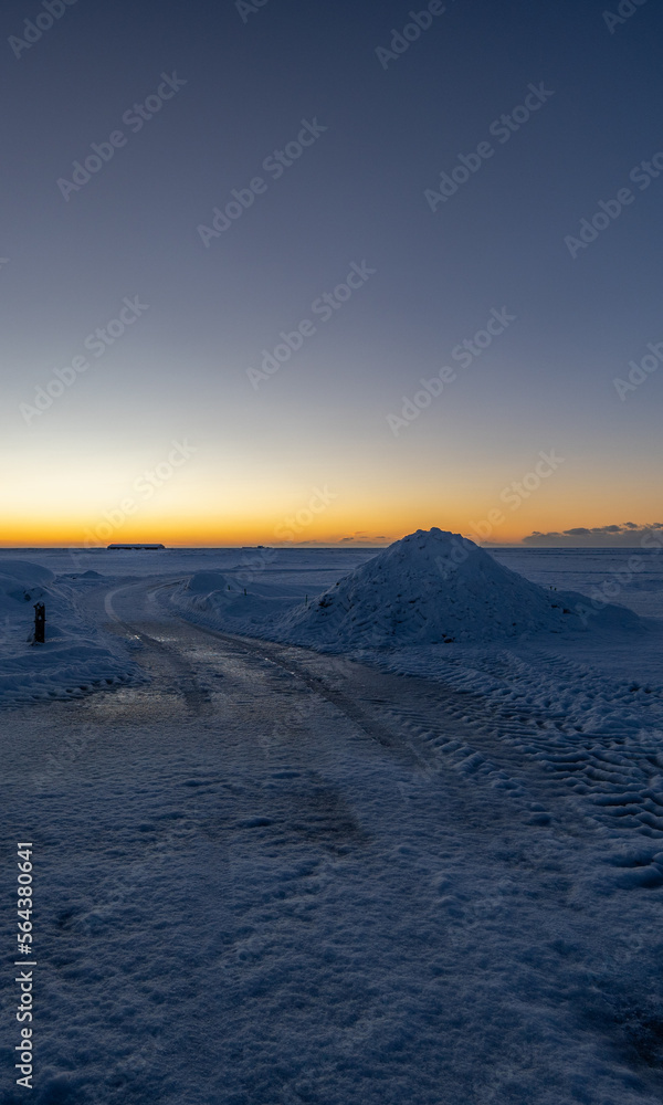 Road and flat snowy landscape in almost darkness with tire tracks going to the horizon with the sun rising in a magical golden Icelandic sunrise.