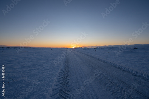 Road and flat snowy landscape in almost darkness with tire tracks going to the horizon with the sun rising in a magical golden Icelandic sunrise. © sirbouman