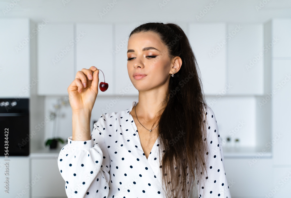 a beautiful European brown-haired girl stands in a white kitchen in a beautiful dress with polka dots and holds a red cherry in her hands and looks not here. Concept kitchen, healthy food, confectione