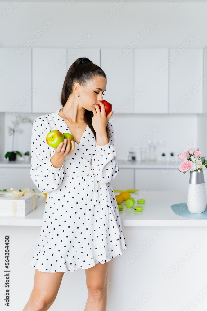 a beautiful european girl stands in a white kitchen in a white polka-dot dress and holds apples in her hands. Concept confectioner, kitchen, equipment, healthy food, nutritionist