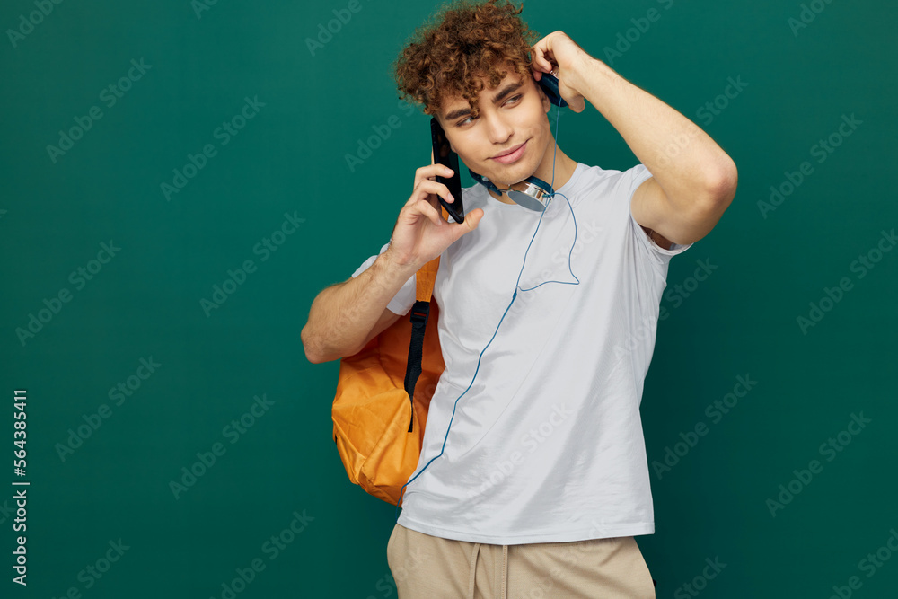 horizontal portrait of a handsome man with curly hair standing on a green background in a gray T-shirt, holding headphones around his neck, talking enthusiastically on the phone