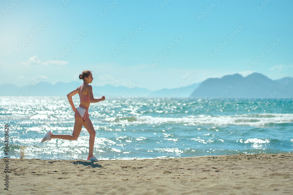 Side view of active sport woman running in morning along the beach. Fit girl jogging on seashore with mountains, cardio workout training outdoors