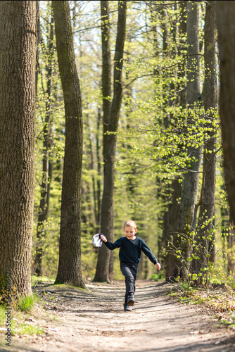 Little boy running hopping along the road in the forest enjoying warm sunny spring weather, holding his hat off in his hand