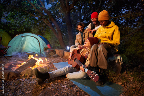 Multiracial colleagues sitting together beside fire stake playing on the guitar discussing and singing. Group of friends with guitar near bonfire and camping tent outdoors overnight. Copy space.