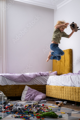 boy having fun, jumping on bed in messy childrens room, kid playing among the many toys at home, a lot of toys and things scattered on floor room, dirty home