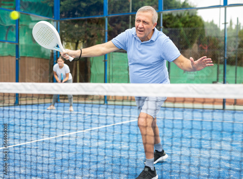 Positive elderly male player serving ball during training padel in court outdoors in spring