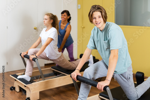 Young boy looking in camera while training pilates in gym. Their trainer hispanic woman helping girl with exercises on pilates reformer in background..