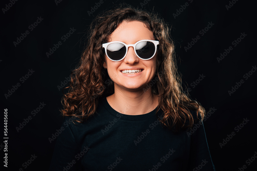 Handsome curly young woman is wide smiling while wearing white sunglasses.