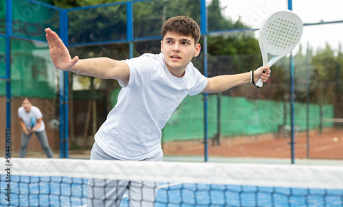 Young European guy playing padel behind the net during match on tennis court in autumn