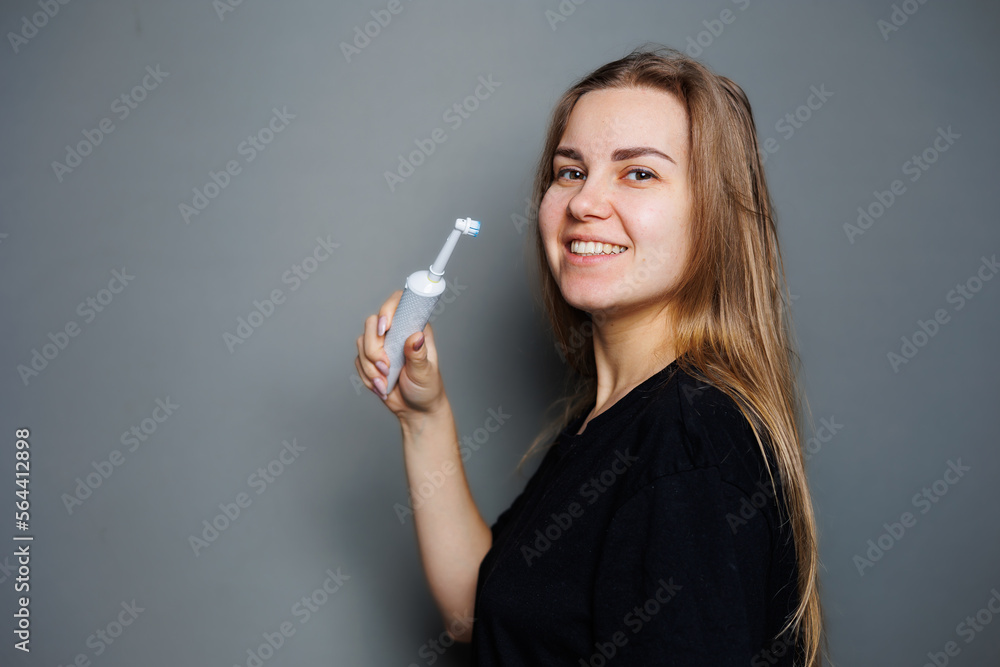 Obraz premium Portrait of a smiling young woman without makeup in a black t-shirt brushing her teeth, mature woman, natural beauty concept. Morning brush for teeth, dental ultrasonic brush.