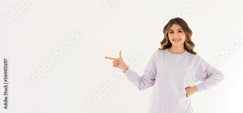Smiling professional woman in purple sweater, pointing finger left at logo and looking confident at camera, standing on white background.
