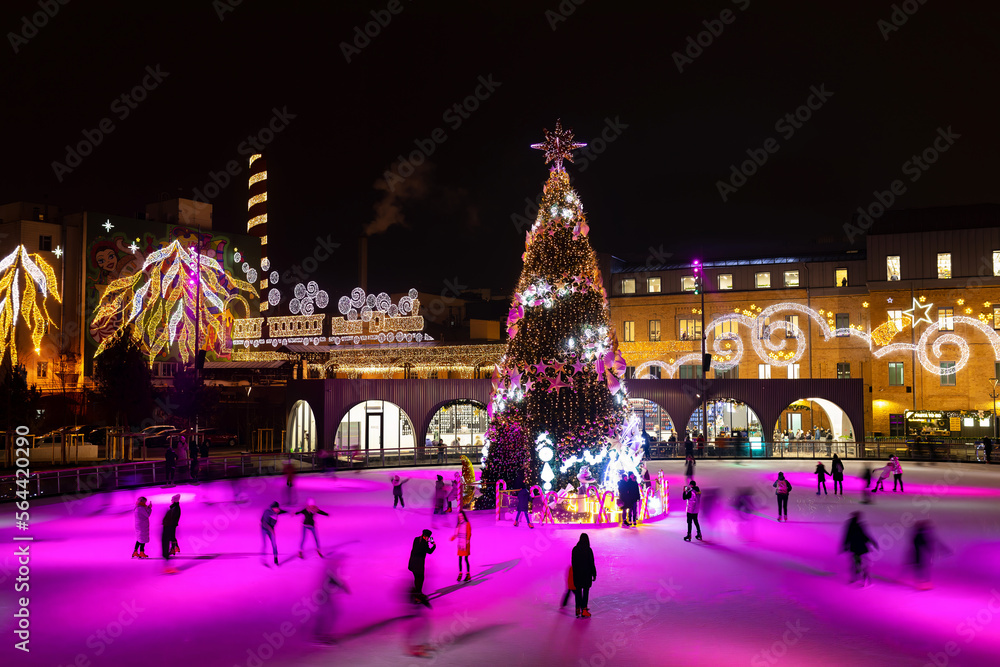 People skate around the Christmas tree at the ice rink, illuminated by pink lights