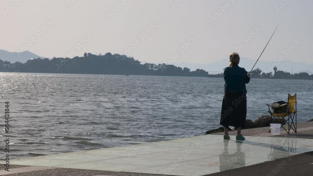 Woman fisherman at the sea bay. An elderly woman with a fishing rod stands on the sea promenade. Woman fisherman at the sea bay. An elderly woman with a fishing rod stands on the sea promenade.