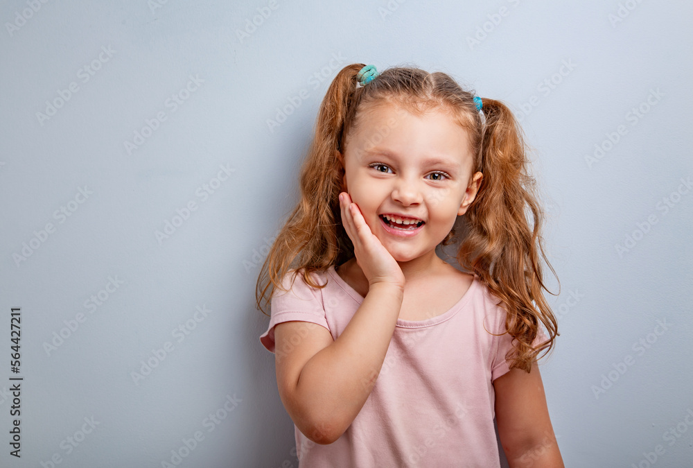 Emotional fun kid girl smiling with hand at face and looking happy with ponytail hairstyle on blue background. Closeup portrait Emotional fun kid girl smiling with hand at face and looking happy with ponytail hairstyle on blue background. Closeup portrait