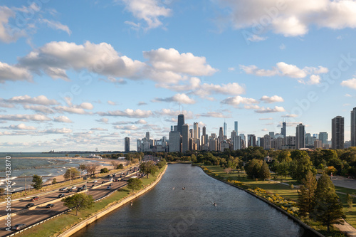 Aerial view of downtown Chicago skyline as a few people use the South Lagoon canal for boating and rowing below on a sunny day with blue sky and fluffy white cumulus clouds above.