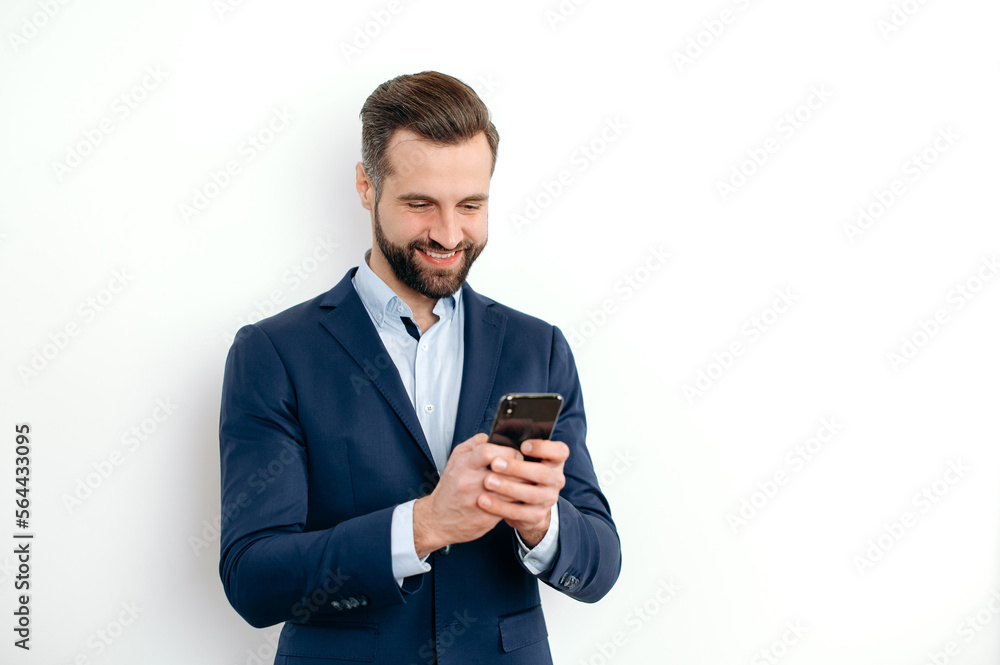 Positive successful caucasian business man in elegant suit, stands on a white isolated background, uses his smartphone, chats online on social networks, looks to the phone, typing text, smiles happily Positive successful caucasian business man in elegant suit, stands on a white isolated background, uses his smartphone, chats online on social networks, looks to the phone, typing text, smiles happily