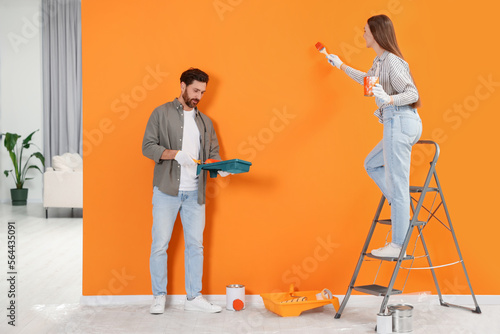 Woman painting orange wall and man holding container with roller indoors. Interior design
