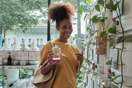 A Black female customer fills liquid soap into recycling jar from reusable containers in a zero-waste and refill store, smiles, and looks at camera, happy shopping in an environment-friendly shop. photo